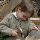 A young child wearing a Halcyon Yarn's Portuguese Fisherman Pullover is focused on hammering a nail into a piece of wood. Surrounded by wooden planks, the scene suggests playful involvement in a family woodworking project. The background includes grass and more wood structures, perfect for this beginner's exploration.