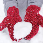 A person is holding a small pile of snow in their red mittens. The background is snowy, creating a wintery scene. The red mittens, which appear to be crafted using the Candide Mitten Pattern by Halcyon Yarn, contrast sharply with the white snow and the light-colored sleeves of the person's jacket.