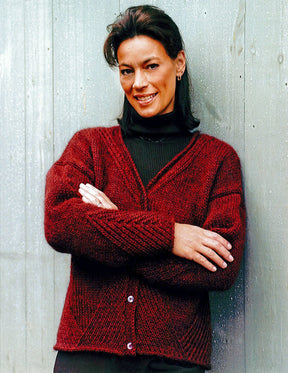 A person stands against a light-colored wooden background, smiling with crossed arms. They are wearing an elegant maroon Diagonal Detail V-Neck Cardigan by Halcyon Yarn over a black turtleneck and have short, dark hair.