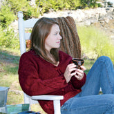 A person with long brown hair is sitting and relaxing on a white wooden chair outdoors, surrounded by greenery. They are wearing the elegant Diagonal Detail V-Neck Cardigan by Halcyon Yarn along with blue jeans, holding a dark-colored mug in both hands. A woven blanket is draped over the back of the chair.