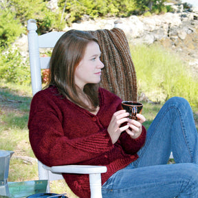 A person with long brown hair, wearing one of their favorite sweaters—a Diagonal Detail V-Neck Cardigan from Halcyon Yarn in red—and blue jeans, sits in a white rocking chair outdoors. They hold a small dark mug. Behind them are trees and rocks, and a braided item is draped over the chair back.