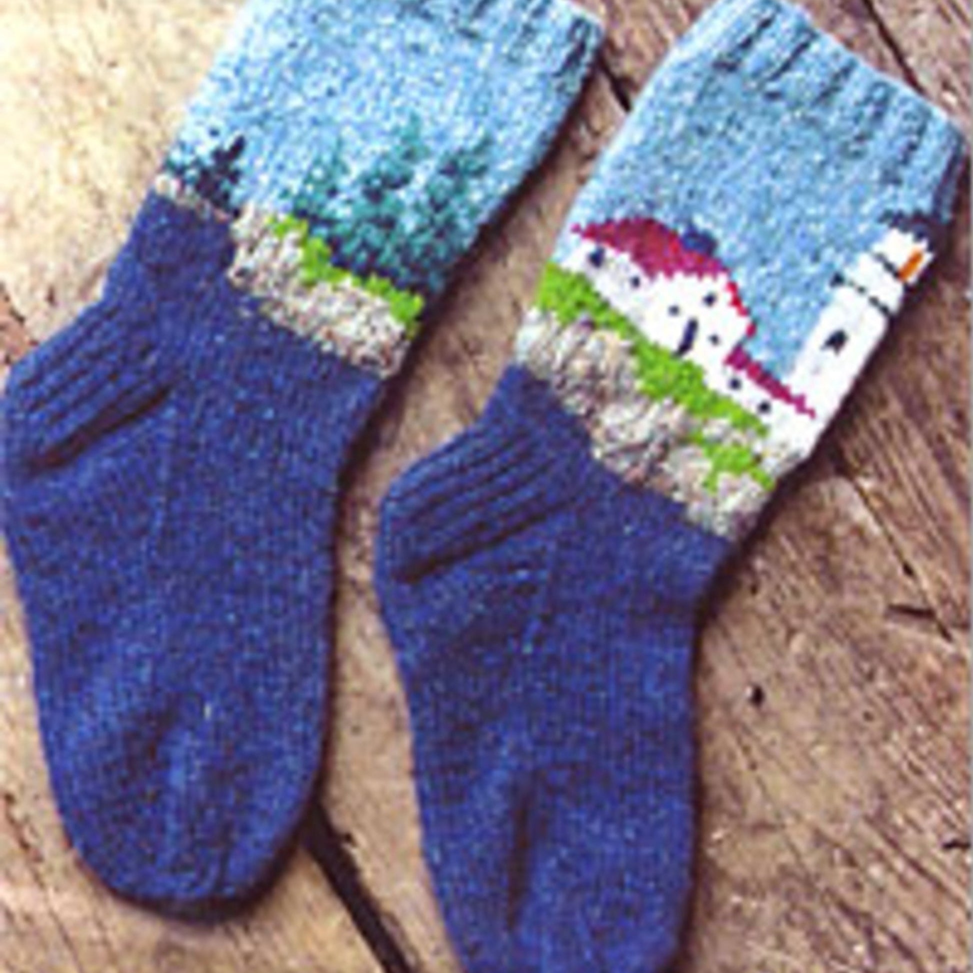 A pair of Lighthouse Socks by Harrisville Designs, showcasing a vivid blue knitted design for adults. These socks feature an intricate lighthouse illustration, complete with a white house that has a red roof, surrounded by greenery and set atop a cliff. The socks are displayed on a wooden surface.