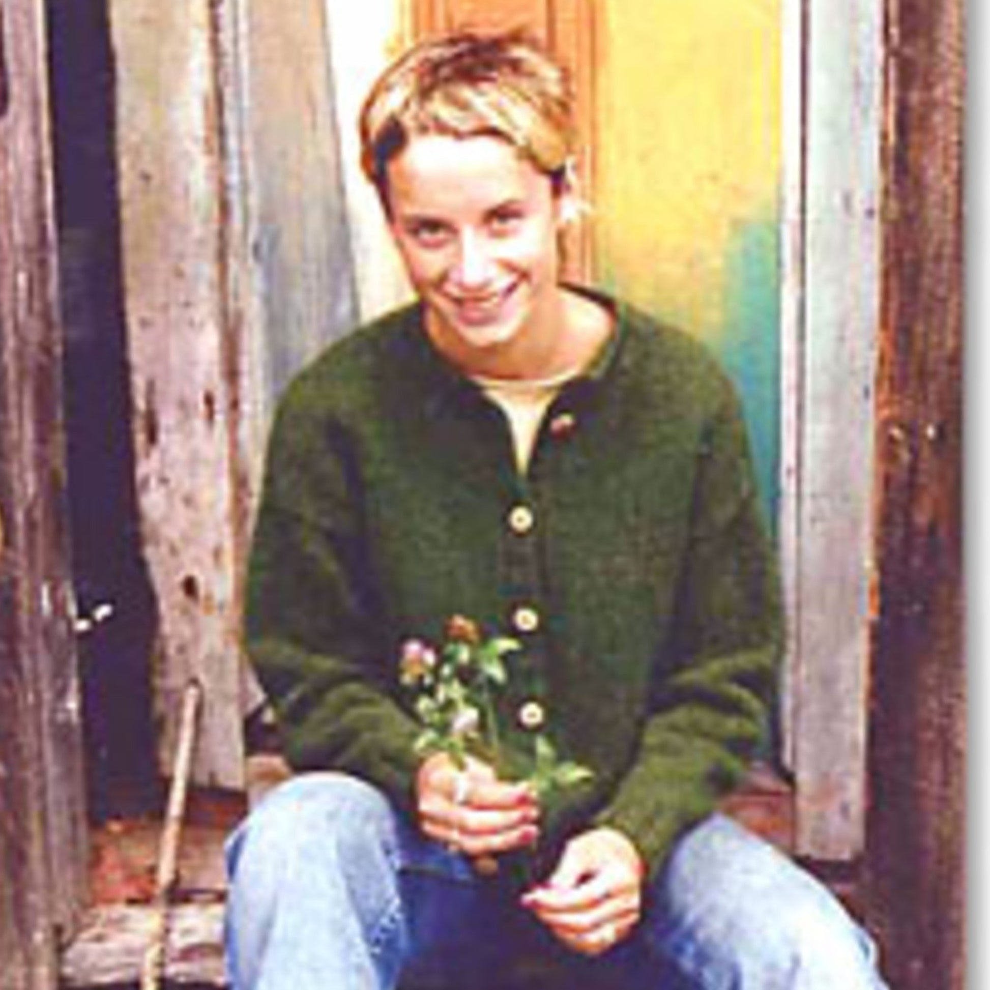 A person with short hair is seated on a rustic wooden step in front of a partially open door. They are wearing a Harrisville Designs Everyday Cardigan in two gauges, paired with blue jeans, and holding a small bundle of flowers in their hands. The background features weathered wooden textures.