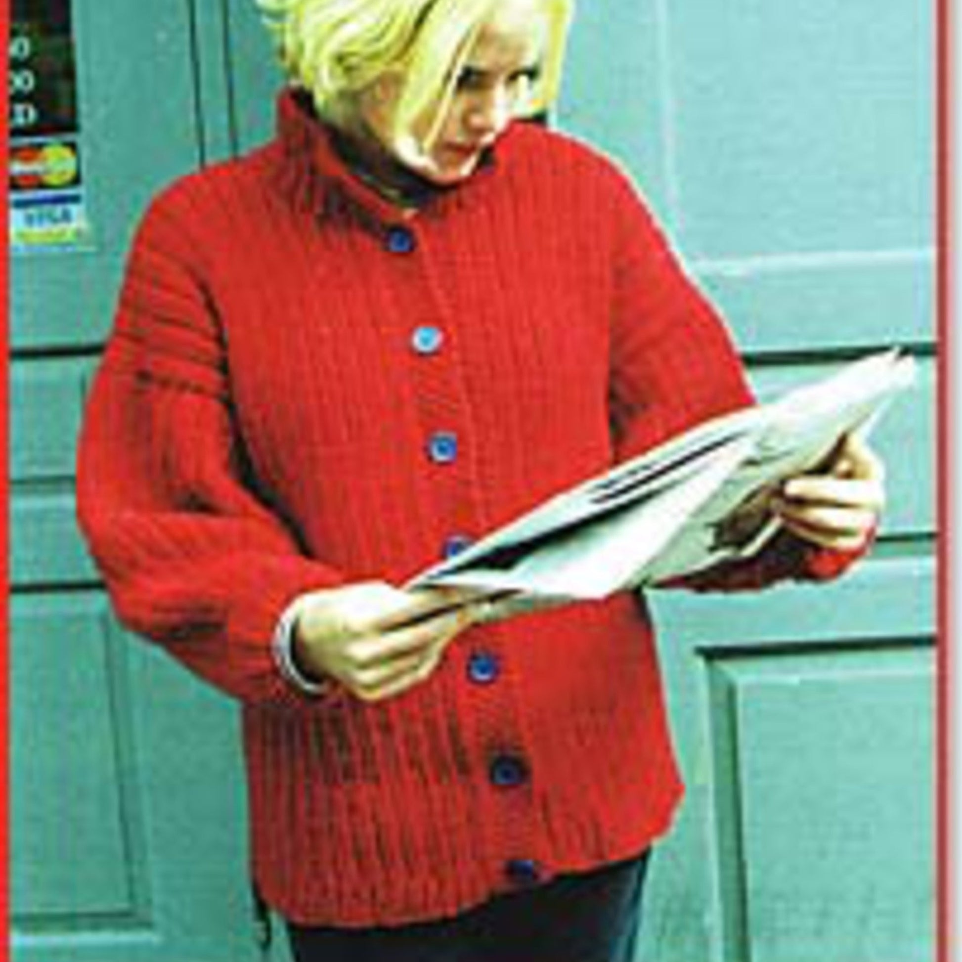 A person with blonde hair wearing a Peace Fleece Ribbed Jacket Sweater from Harrisville Designs is standing outdoors against a blue door, reading a newspaper.