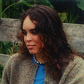 A woman with wavy brown hair and a necklace of large beads smiles gently. She is wearing the Sassy Cardigan Sweater from Harrisville Designs over a blue shirt, giving her an elegant uptown look as she sits outdoors on a wooden bench, with greenery in the background.