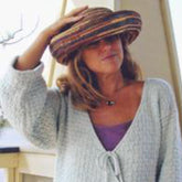 A woman with wavy blonde hair, wearing a Bryson Distributing Inc.'s Monterey Wrap Cardigan in light gray featuring a Tiny Bowknot pattern over a purple top, adjusts a large, colorful straw hat on her head. She stands in front of a light background, possibly a porch or deck, with part of a plant visible to the left.