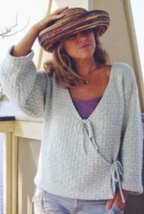 A woman wearing a wide-brimmed colorful hat and a relaxed Monterey Wrap Cardigan by Bryson Distributing, Inc. over a purple top is standing and touching the brim of her hat with one hand. The background appears to be an outdoor setting, possibly a porch or terrace.