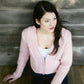 A woman with long dark hair, wearing the Bryson Distributing, Inc. Newport Jacket in light pink and a white top, sits on wooden floorboards against a wooden wall. She gazes to the left with a serious expression, her hair loose over one shoulder. The setting is rustic and simple.