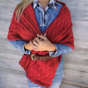 A person wearing the Magnolia Wrap from Bryson Distributing, Inc. draped over a denim shirt with a plaid collar peeking out, cinched at the waist with a brown braided belt. The individual has crossed hands in front and is standing against a wooden background. Only the lower face and chest are visible, giving the wrap an elegant touch.