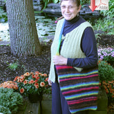 A smiling person wearing glasses, a knitted vest, and carrying a colorful Knottogo Bag by Halcyon Yarn with an adjustable strap, stands near a pond surrounded by foliage and potted flowers. An arrow with the text "Adobe" points to the person from the top right corner of the image.