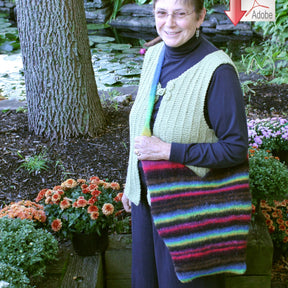 A smiling person stands outdoors, wearing glasses and a knitted vest, holding the colorful, striped KnotToGo Bag with an adjustable strap. They are surrounded by flowers and greenery with a pond in the background.