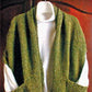 A Reader's Wrap by Lisa's Knits, featuring a textured pattern in green with a wide collar and front pockets, is draped over a white long-sleeved top. Both garments are hanging on a hanger against a wooden background. This shawl is perfect for when you feel chilled, making it an ideal choice to snuggle up in while reading.