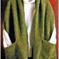 A green, textured knit cardigan, known as the Reader's Wrap by Lisa's Knits, hangs gracefully draped over a white hanger. This cozy piece, ideal for those chilly moments, is showcased against a wooden background and seems to be layered over a white shirt. Two large front pockets add both style and functionality to this elegant cardigan.