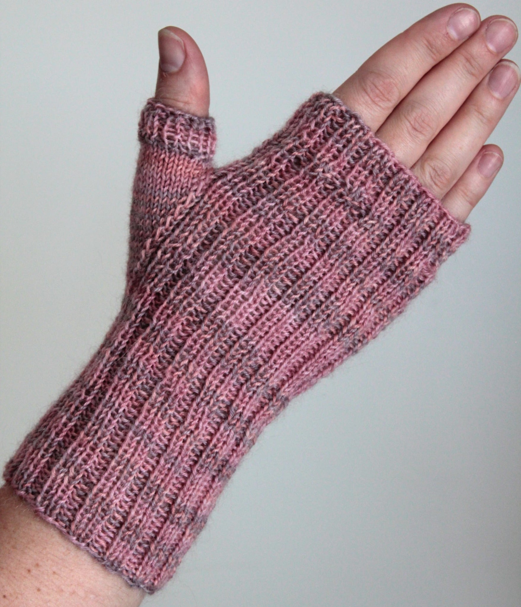 A hand wearing Lisa's Knits' Fingerless Gloves and Mitts, Fingering Weight, made of pink and gray variegated yarn. The glove features a ribbed pattern with an open thumb hole, leaving the fingers free. Crafted from high-quality fingering weight yarn, these gloves are showcased against a plain light-colored background.