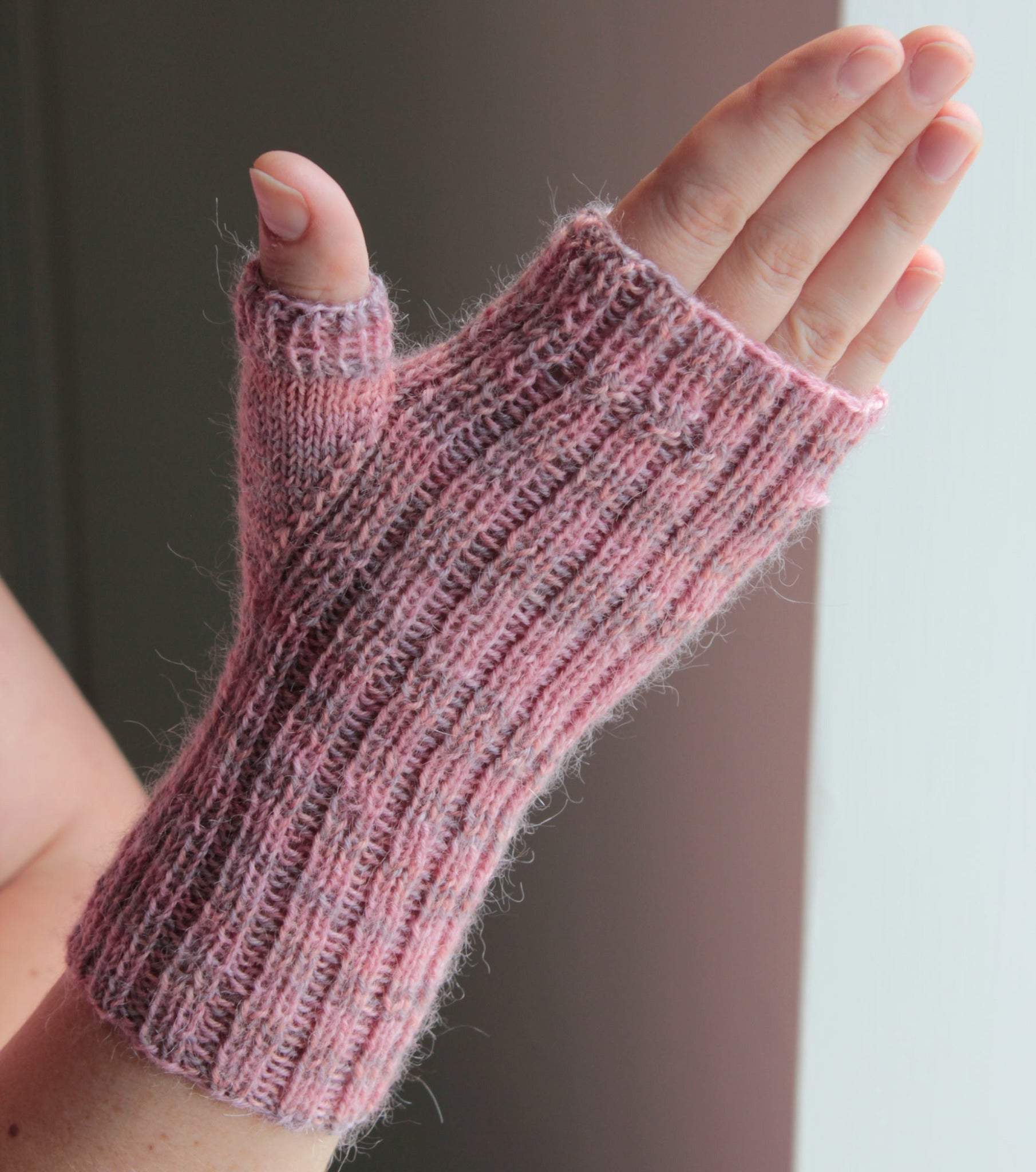 A hand wearing Lisa's Knits' Fingerless Gloves and Mitts, Fingering Weight, in pink is shown against a neutral background. The mitt features a ribbed texture and covers only the thumb, leaving the fingers free.