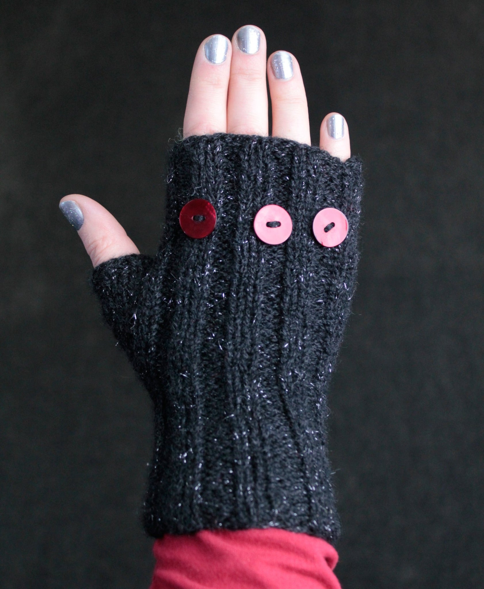 A hand wearing a black Lisa's Knits Fingerless Glove, adorned with three red buttons on the back. The hand's nails are painted with silver polish, and the background is dark. This winter accessory covers the wrist and part of the forearm, providing stylish warmth.