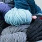 Close-up of several skeins of self-patterning yarn in varying colors, including purple, teal, black, gray, and cream. A person wearing a pair of Lisa's Knits "Fingerless Gloves and Mitts 5 sts/1"" adorned with red buttons is arranging the yarn on a flat surface.