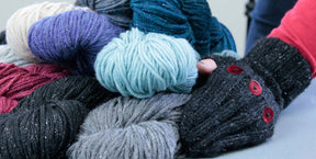 Close-up of several skeins of self-patterning yarn in varying colors, including purple, teal, black, gray, and cream. A person wearing a pair of Lisa's Knits "Fingerless Gloves and Mitts 5 sts/1"" adorned with red buttons is arranging the yarn on a flat surface.