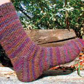 A close-up of a person's foot wearing a vibrant sock from Lisa's Knits, crafted with the Basic Toe-Up Sock 6 st/1" pattern. The sock showcases a stunning blend of red, purple, and brown hues in a striped design. The background features a wooden log and green foliage.