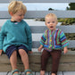 Two young children are sitting on a wooden bench by the waterfront. The older child, dressed in a blue sweater and dark shorts, looks down at the younger child with a smile. The younger child is wearing Schoolhouse Press's Elizabeth Zimmermann's A-B-C Surprise Jacket and brown pants. In the background, you can see water and greenery.