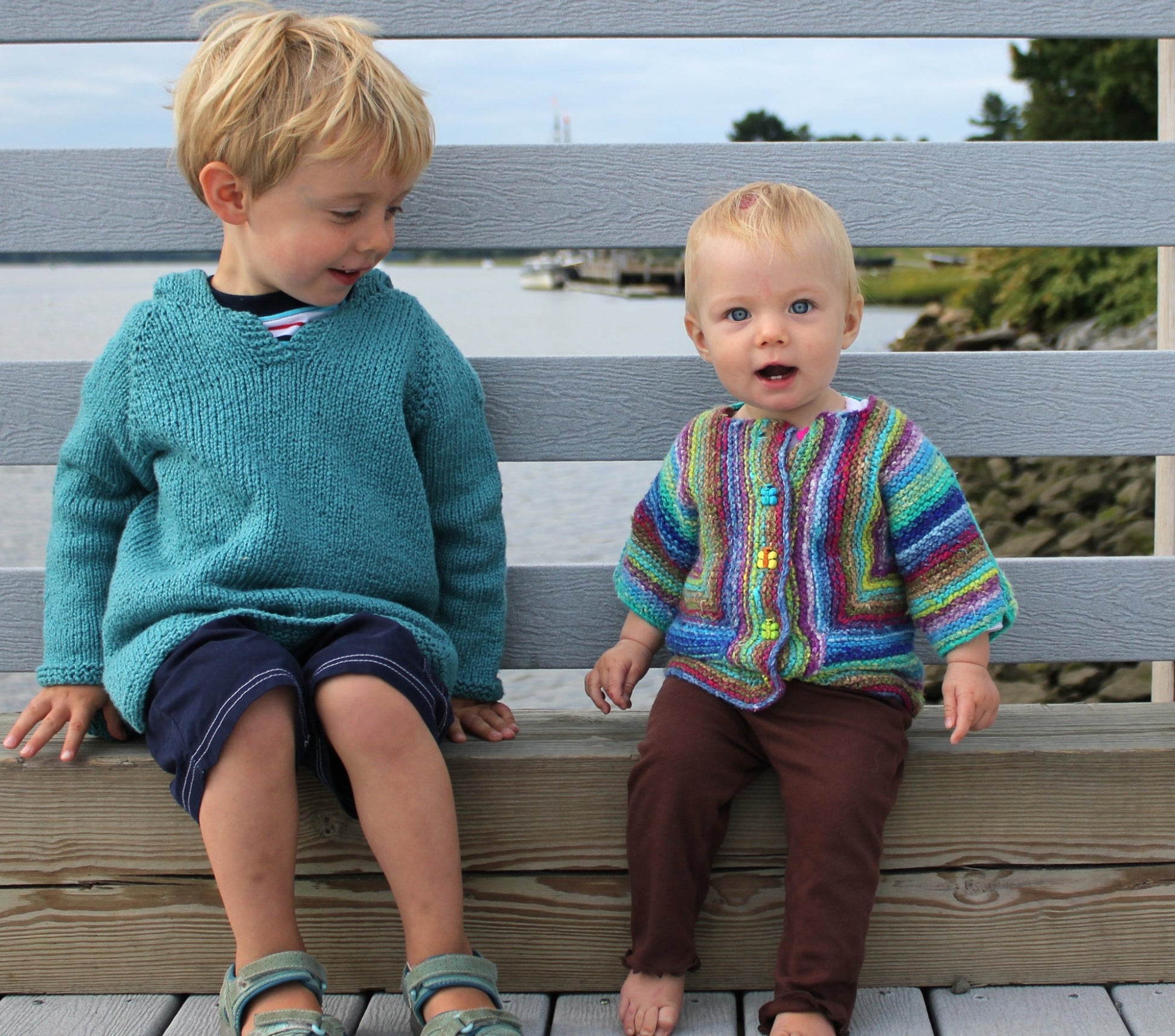 Two young children are sitting on a wooden bench by the waterfront. The older child, dressed in a blue sweater and dark shorts, looks down at the younger child with a smile. The younger child is wearing Schoolhouse Press's Elizabeth Zimmermann's A-B-C Surprise Jacket and brown pants. In the background, you can see water and greenery.