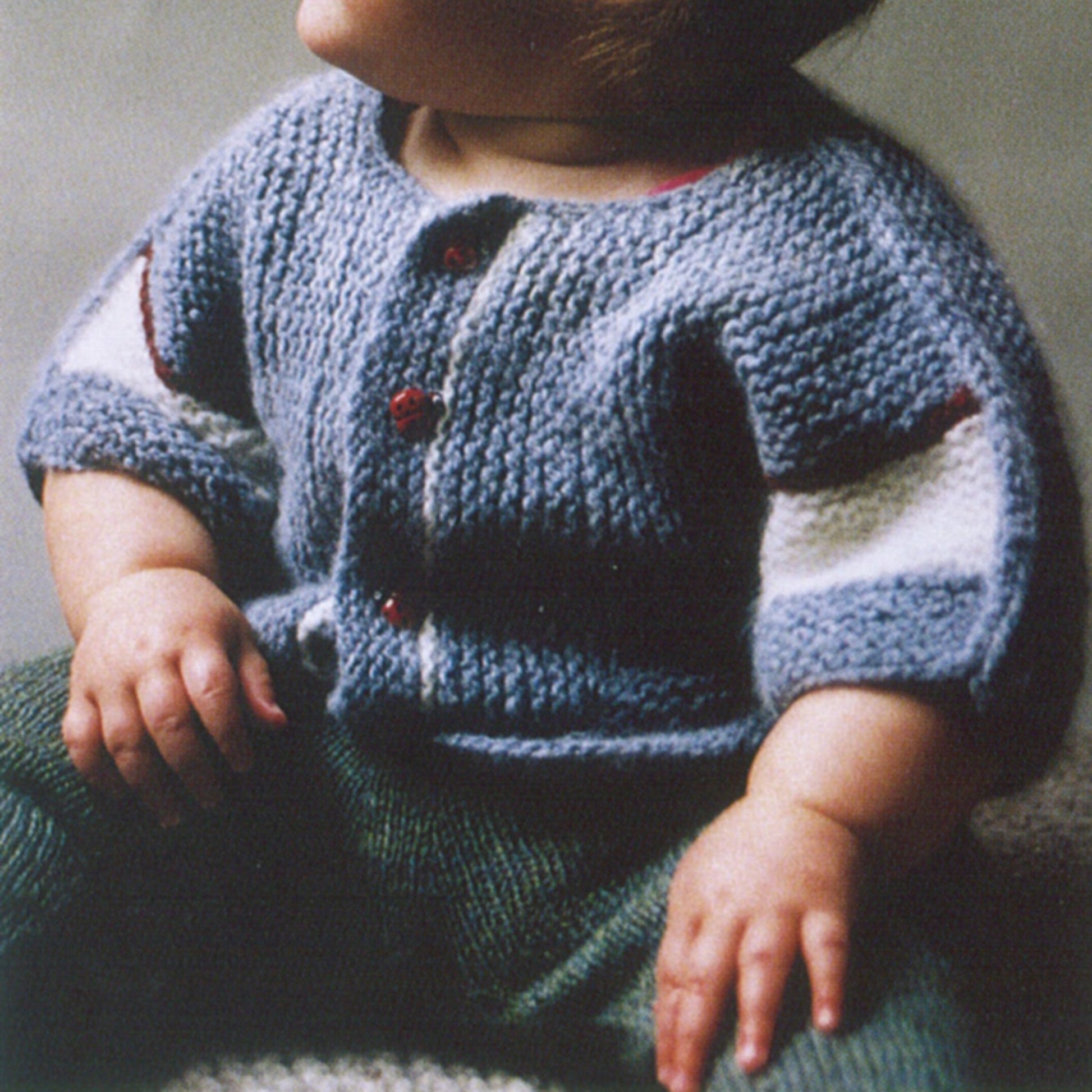 A baby wearing the Elizabeth Zimmermann's A-B-C Surprise Jacket by Schoolhouse Press is seen sitting with hands resting on their lap. The blue knitted sweater, adorned with red buttons and a charming garter stitch pattern complete with white patches on the sleeves, beautifully complements the green pants. The softly lit background emphasizes the intricate knitting pattern of this delightful knitwear.