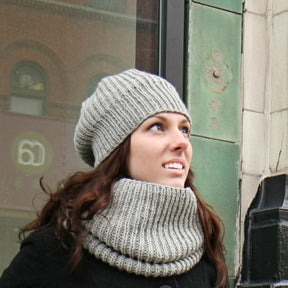 A woman with long brunette hair is wearing a grey knit hat with a ribbed band and matching scarf from Chic Knits' Super Cupcake collection. She is looking upwards and standing next to a building with green and beige tiles. A blurred storefront window and brick building are visible in the background, perfectly complementing the Super Cupcake series.