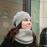 A woman with long brown hair, wearing Chic Knits' "Super Cupcake" hat made of worsted weight yarn along with a matching scarf, looks up while standing outside next to a building adorned with green tiles and a display window. Dressed in a black coat, the background features part of a brick wall with reflective surfaces.