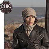 An individual clad in a knitted cap, scarf, and leather jacket from the Elisbeth Collection stands outside near a beach on a chilly day. The person holds a warm drink and is framed by a wooden post. The upper left corner features the "Chic Knits" logo.