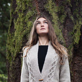 A woman with long brown hair, wearing the Chic Knits' Tauriel Cardigan, a beige knit with chain-cable detailing, over a black top stands in front of a large tree trunk with moss. She gazes upward with a contemplative expression against a backdrop filled with greenery.