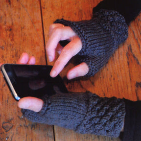 Hands adorned with the cozy Blackberry Mitts Pattern from Swans Island Yarns, ideal for texting or using a smartphone on a wooden table.