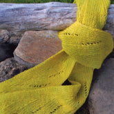 The Shot of Spring Scarf by Swans Island Yarns, with its light and airy yellow lacy pattern, is draped over a weathered wooden railing against a backdrop of rocks and blurred greenery.