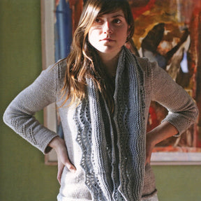 A young woman with long brown hair stands in front of a colorful painting, wearing a gray knitted sweater and the Anna Cowl from Swans Island Yarns. She has her hands on her hips and looks slightly towards the left. The background is green.