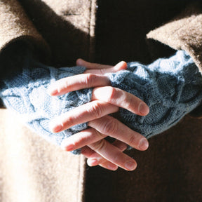 Close-up of a person in a brown coat with hands clasped, wearing Swans Island Yarns' Earl Grey Mitts. The image focuses on the hands and a part of the coat, capturing the texture of the intertwining cables and fabric.