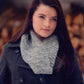 A young woman with long dark hair is wearing a dark gray peacoat and the Ava Cowl from Swans Island Yarns, styled as a light gray knitted scarf. She gazes slightly to the side with a calm expression. The softly blurred background suggests an outdoor setting, evoking a sophisticated alternative vibe.