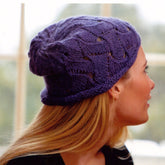 A person with long blonde hair sports the Brynn Hat, a slouchy purple knit beanie crafted with a textured, lacy pattern by Swans Island Yarns, perfectly capturing the boho-chic vibe. Indoors, they're seated facing a window with a softly blurred outdoor view.