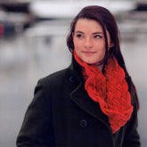 A young woman with long, dark hair, wearing a dark green coat and the chunky red Entwined Cowl by Swans Island Yarns, stands outdoors on a cloudy day, looking off to the side with a slight smile. The background is out of focus, creating a soft, serene atmosphere perfect for winter days.