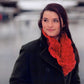 Dressed warmly for the chilly winter day, a person stands outdoors in a dark coat, showcasing the vibrant red Entwined Cowl by Swans Island Yarns, which adds a touch of brightness to their ensemble as they gaze to the side with a slight smile. The blurred background enhances the wintry ambiance.