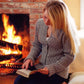 A person with long blonde hair, wearing the Eleanor Cardigan by Swans Island Yarns, is sitting in front of a roaring fireplace. They are focused on reading an open book, creating a cozy and warm atmosphere.