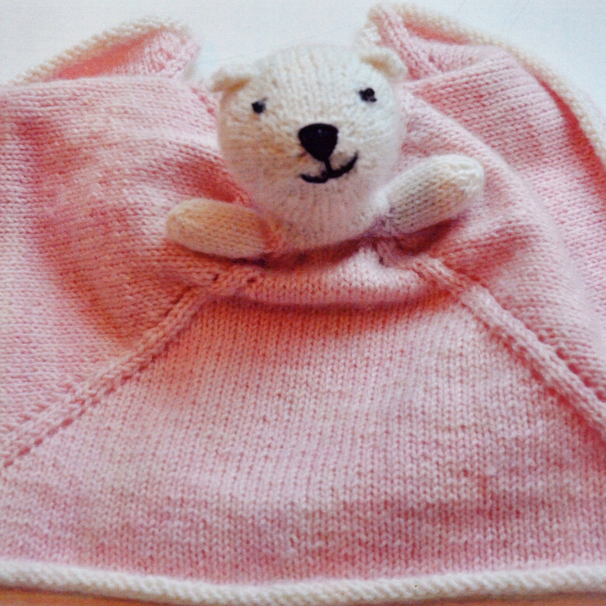 Introducing the Ballerina Bear by Swans Island Yarns: a charming knitted pink blanket showcasing the adorable head and tiny hands of a white bear plush toy emerging from the center. The lovable bear features black stitched eyes and nose, while ribbed edges give the blanket a neat finish.