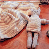 A knitted toy cat with a smiling face, gray and beige stripes, from the "Cat and Hat" collection by Swans Island Yarns, lies on a wooden surface next to a matching knit hat. Nearby, there is a partially knitted fabric piece still on the knitting needles. Yarn and knitting accessories, including Swans Island Organic Merino Ecowash, are also visible in the background.