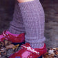 A child wears knit knee-high leg warmers, designed with the Aspen Leg Warmers Pattern by Swans Island Yarns, in a soft lavender hue adorned with a delicate cable pattern. These are beautifully paired with shiny red shoes featuring decorative trim and blue flower accents. Fallen leaves are scattered on the ground around their feet.