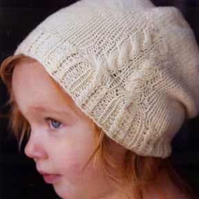 A young child with reddish hair is wearing a cream-colored hat made from the White Pines Hat Pattern by Swans Island Yarns, featuring a ribbed brim. The child gazes off to the side with a neutral expression, set against a dark background that highlights the intricate texture of the hat and the child's profile.