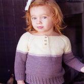 A young child with red hair is standing indoors, wearing a Swans Island Yarns Juniper Sweater Pattern that showcases a lovely cream and mauve color block design with three buttons on the front. Their hair is adorned with a white bow, adding charm to an ensemble that would be perfect for any children's wardrobe.