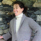 A woman with dark hair tied back is smiling and leaning against a stone wall. She is wearing a Bryson Distributing, Inc.'s light gray Kimono Cardigan over a white turtleneck sweater, showcasing her knitting skills. The stone wall behind her consists of variously sized rocks.