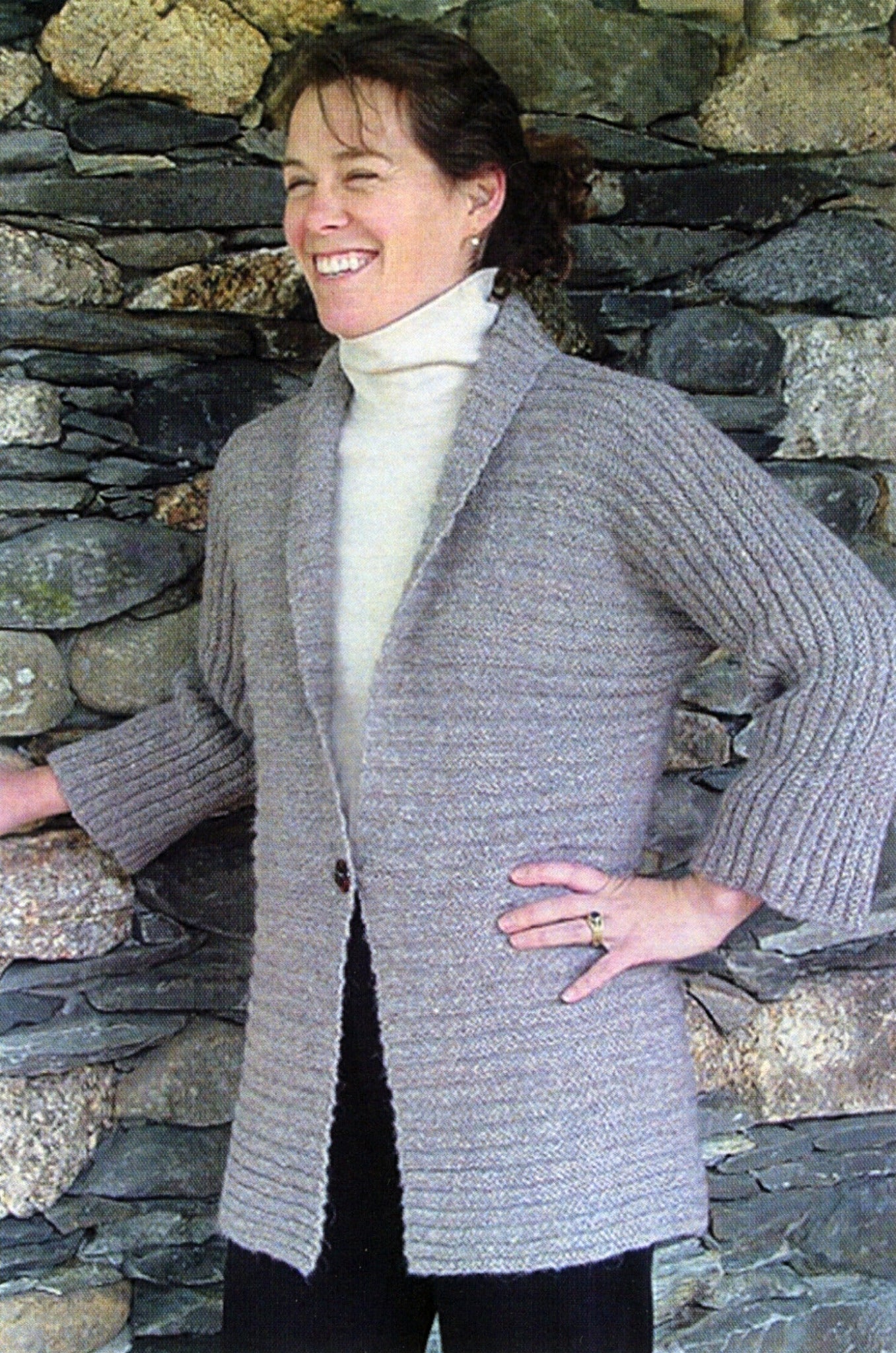 A person is standing in front of a stone wall, smiling. They are wearing a light gray Kimono Cardigan with wide sleeves from Bryson Distributing, Inc. over a white turtleneck sweater and black pants. Their hair is tied back, and they have one hand on their hip and the other resting on the wall.