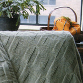 A cozy Reversible Afghan from Bryson Distributing, Inc., in light gray with a knit pattern, is draped over a chair. Behind it, there is a basket filled with colorful fabric and a leafy plant. The scene, featuring rich textures and reversible designs, is set near a window with a view of a brick building outside.