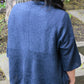 A person with long dark hair wearing the Never Not Knitting Knitbot Wispy Cardi, a loose-fitting, dark blue knit sweater with an open front design, is standing outdoors with their back to the camera. The background includes a tree trunk and stone steps.