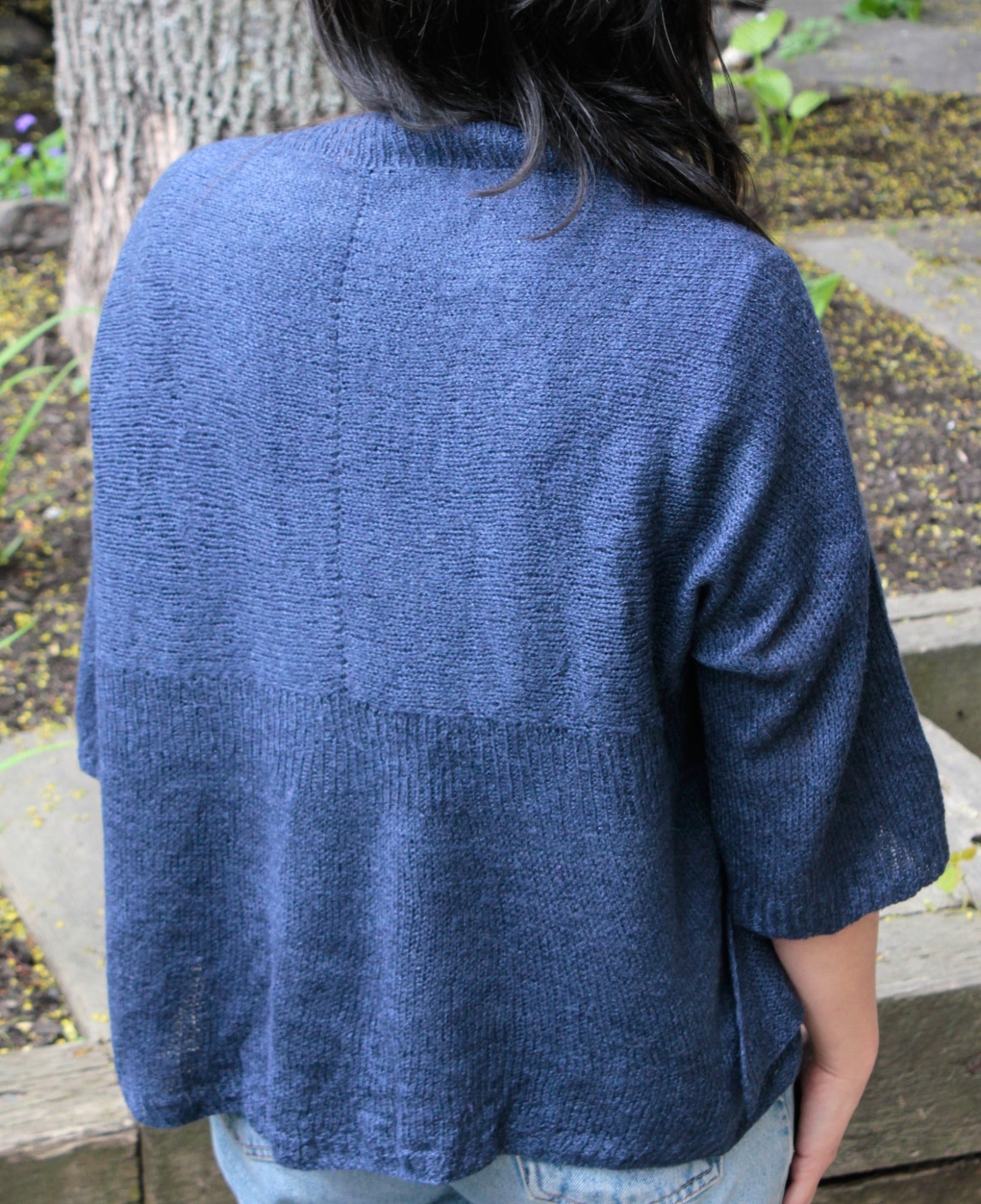 The image shows a person with long dark hair standing outdoors, facing away from the camera. They are wearing a loose-fitting Knitbot Wispy Cardi by Never Not Knitting, which has elbow-length sleeves and is in a blue knit. They are also wearing light denim jeans. The background includes a tree trunk and some leafy plants.