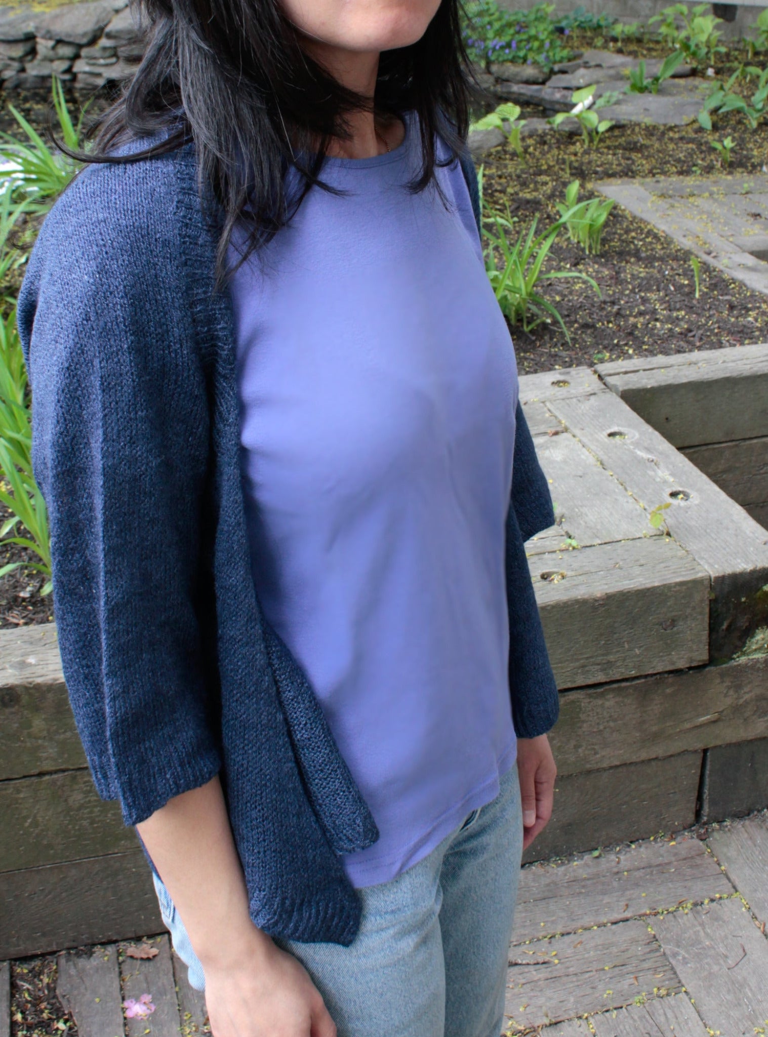 A person wearing a blue shirt and a Knitbot Wispy Cardi, a dark navy lightweight cardigan by Never Not Knitting, is standing outdoors. The background features stone steps and garden beds with green plants. The person is visible from the neck down, with dark hair falling over their shoulders in an open front design.